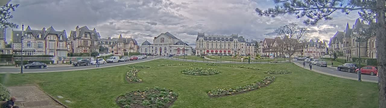 A grassy town square with flowerbeds, surrounded by several ornate buildings under a cloudy sky.