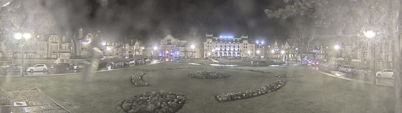 A nighttime scene of a town square with several buildings, cars parked along the sides, and landscaped flowerbeds in the center, under rainy conditions.