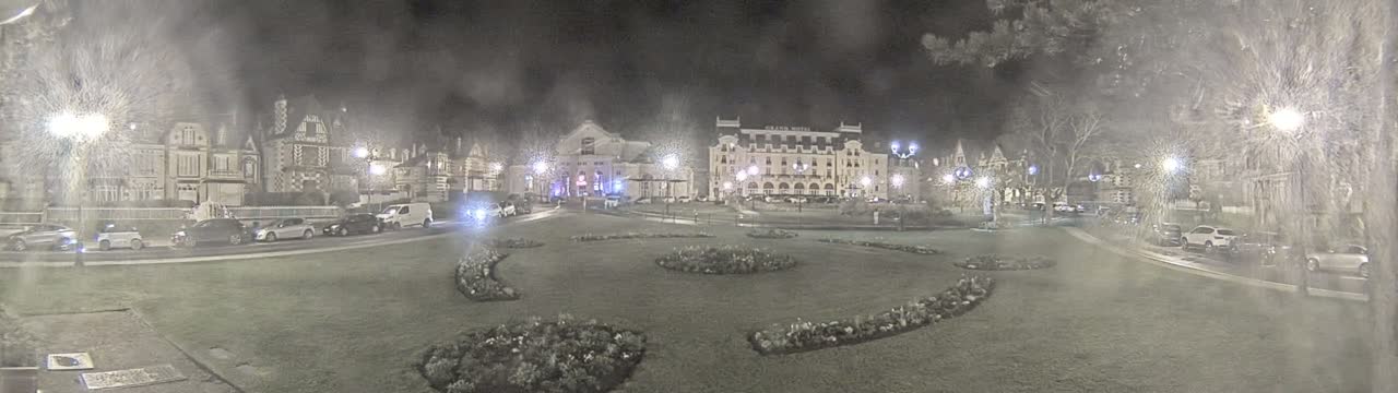At night, under a hazy sky, cars are parked along the edges of a grassy town square containing several flowerbeds, in front of a row of buildings including a large hotel.