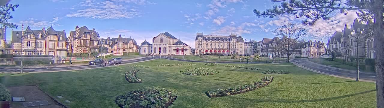 A grassy park with decorative flowerbeds sits in front of a row of ornate buildings under a partly cloudy sky.