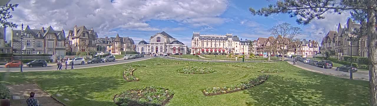 A grassy park with flowerbeds is situated in front of a row of multi-story buildings on a partly sunny day.