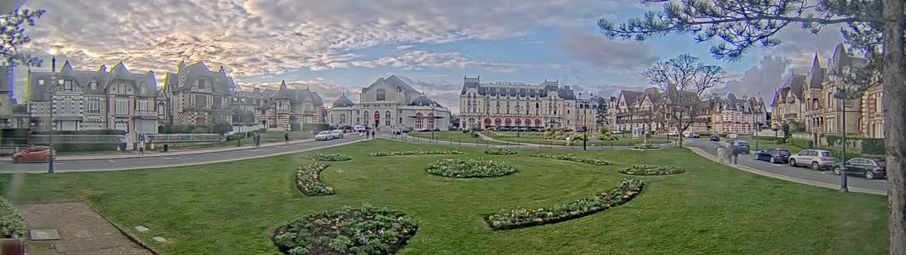 A grassy square with flowerbeds is in front of several large buildings under a partly cloudy sky.