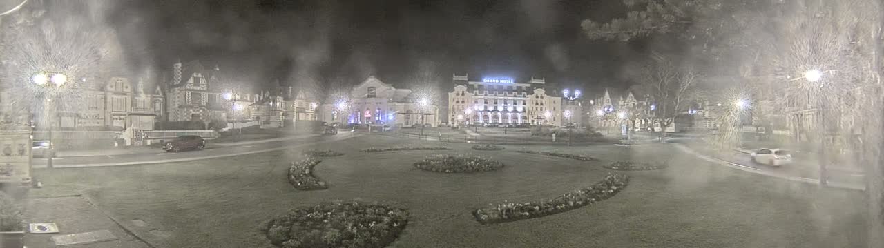 A snowy night scene shows a town square with several buildings, including a large hotel, and landscaped areas with flowerbeds, lit by streetlights.