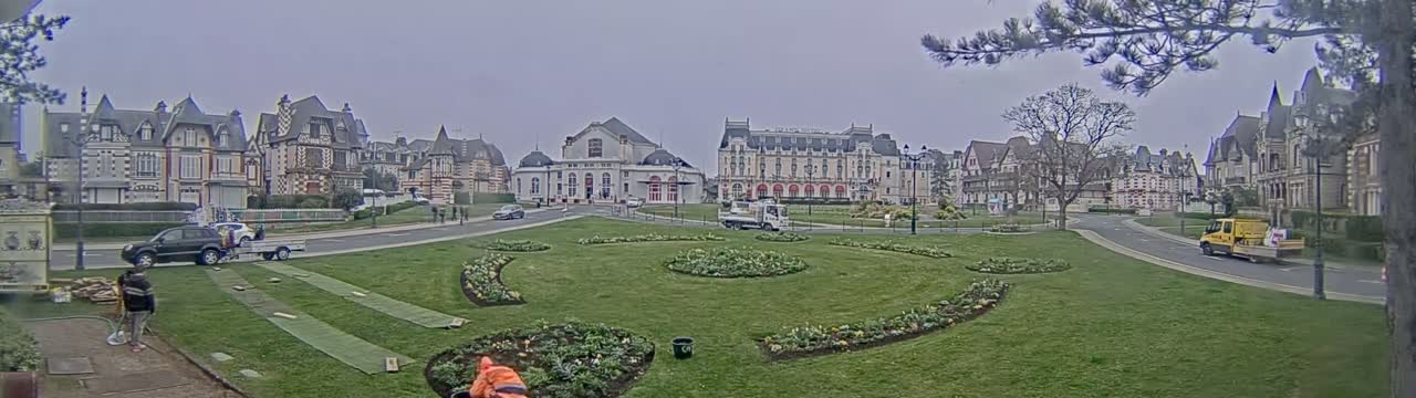 A grassy town square with flowerbeds is surrounded by several large, ornate buildings under an overcast sky.