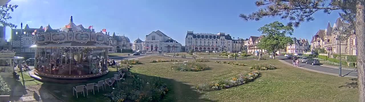 A carousel sits in a grassy town square, surrounded by several buildings on a sunny day.