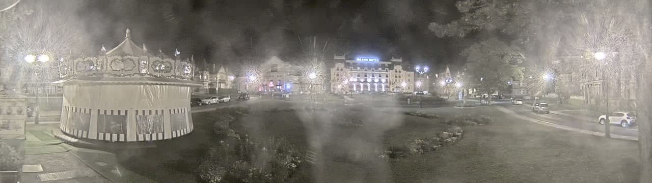 A carousel sits in a snowy nighttime town square in front of several buildings, some cars parked nearby.