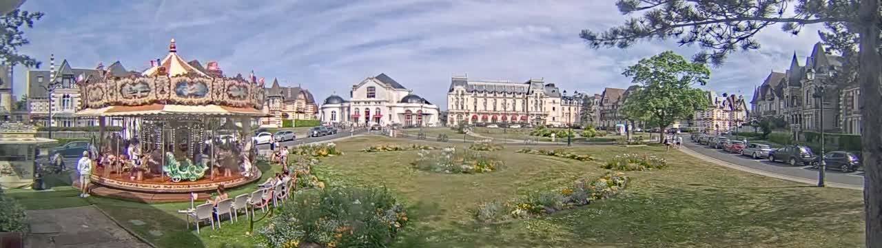 A carousel sits in a town square on a partly cloudy day, surrounded by buildings and a grassy area with flowerbeds.