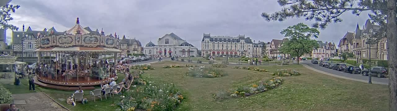 A carousel is in a town square with several buildings, parked cars, and people on an overcast day.