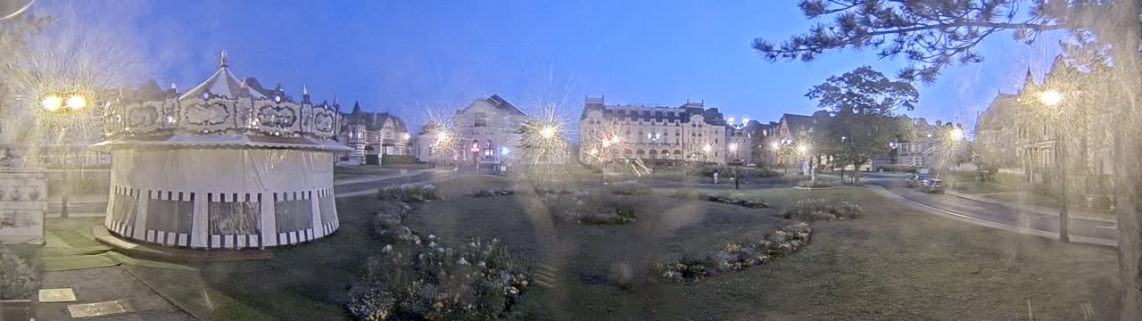 A carousel sits in a park at twilight, near several large buildings under a slightly hazy sky.