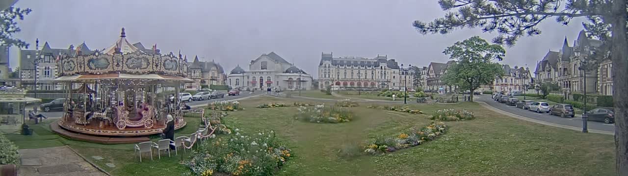 A carousel sits in a grassy town square near several buildings, under an overcast sky.