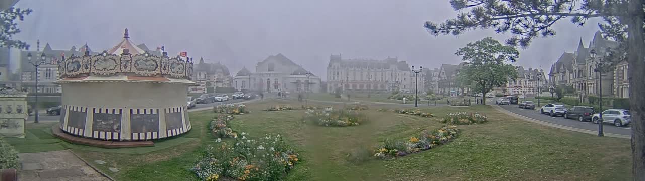 A carousel sits in a grassy town square surrounded by buildings and flowerbeds on a foggy day.