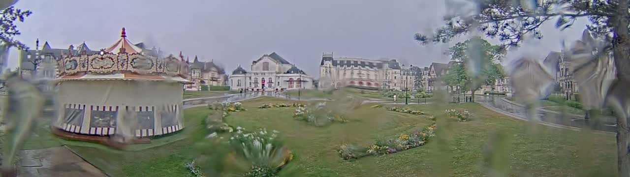 A carousel sits in a grassy park in front of several buildings on a rainy day.