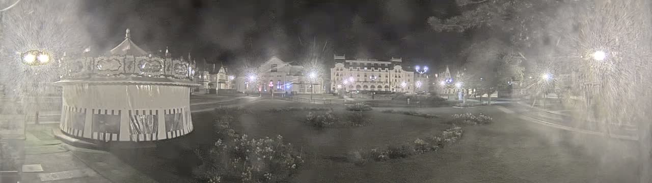 A carousel sits in a snow-covered park at night, with several buildings, including a large hotel, lit up in the background.
