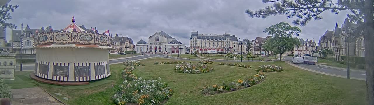 A carousel sits in a grassy park, in front of several large buildings, under an overcast sky.