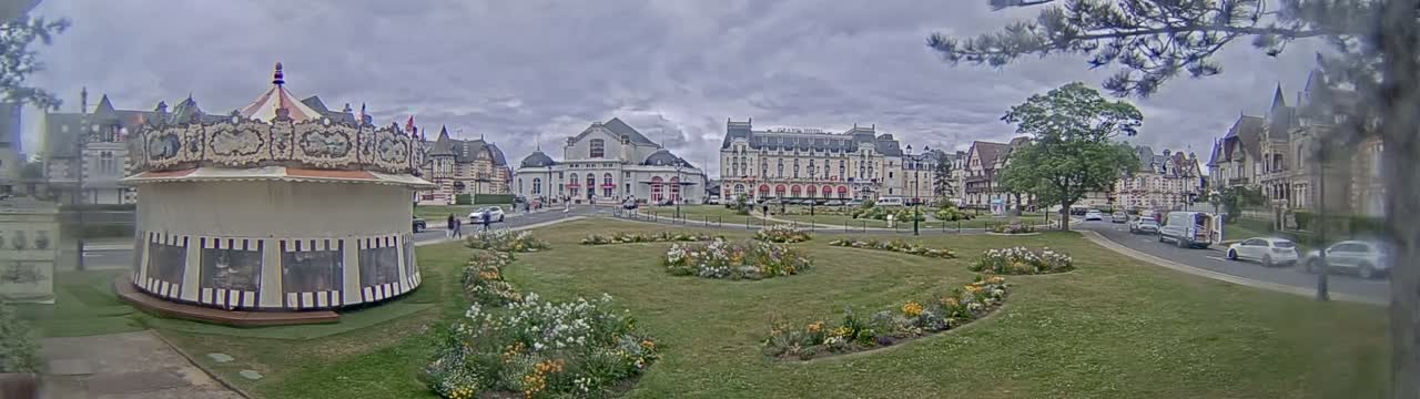 A carousel sits in a grassy town square bordered by flowerbeds, in front of several large buildings under an overcast sky.