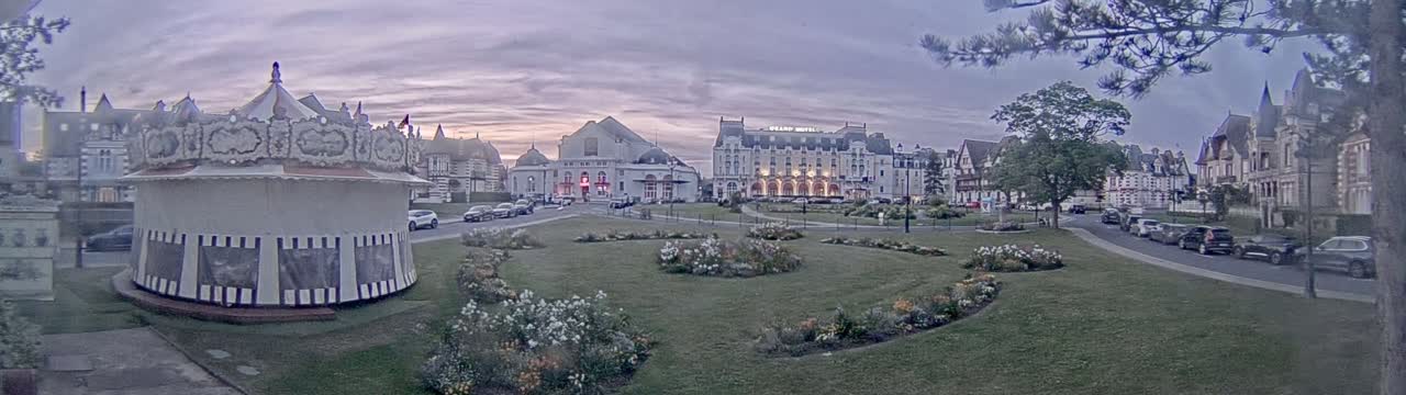 A carousel sits in a park next to several buildings, including a large hotel, under a twilight sky.