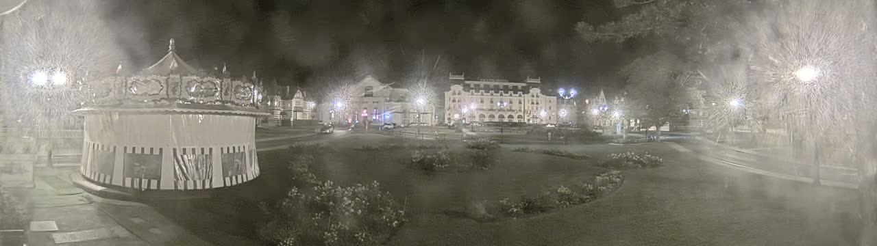 A carousel sits in a snow-covered town square at night, near a large hotel and other buildings illuminated by streetlights.