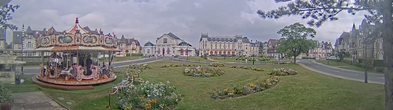 A carousel sits in a town square, surrounded by flowerbeds and several large buildings under a cloudy sky.