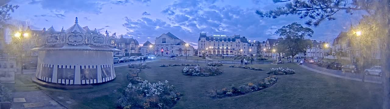 A carousel sits in a twilight town square beside a grassy area with flowerbeds, in front of a row of buildings including a large hotel.