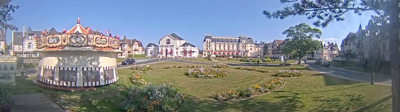 A carousel sits in a grassy town square on a sunny day, with several large buildings and flowering gardens in the background.