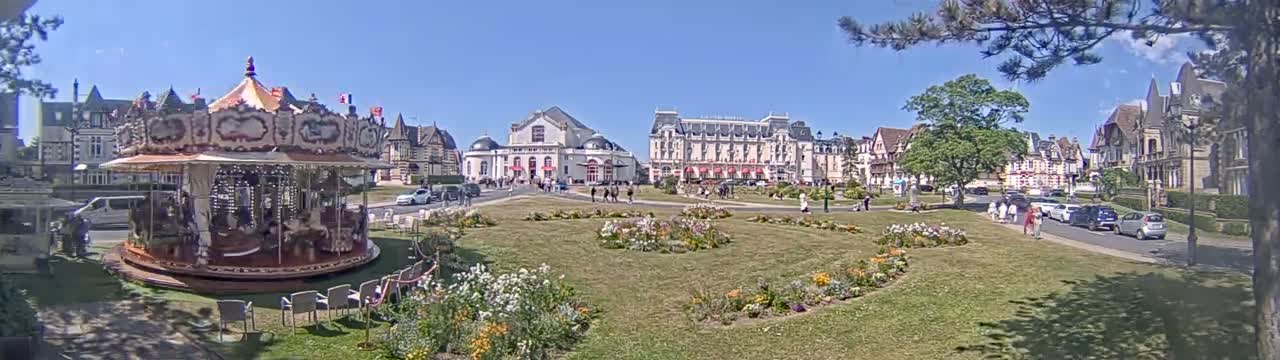 A carousel sits in a grassy town square on a sunny day, surrounded by several buildings and people, with cars parked along a nearby street.
