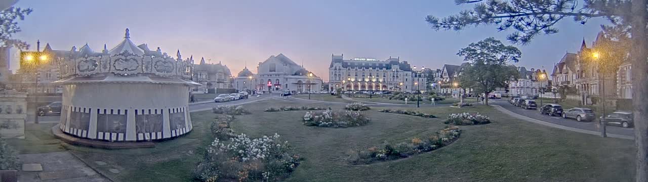 A carousel sits in a town square at dusk, surrounded by several buildings, lush green lawns, and flowerbeds, with cars parked along a nearby street.