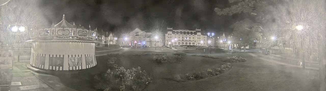 A carousel sits in a snow-covered park at night, near a large hotel and other buildings, under bright streetlights.