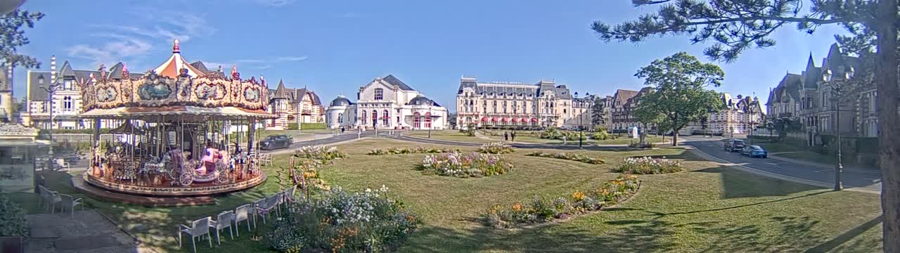A carousel sits in a grassy town square with several large buildings and flower gardens, under a sunny sky.