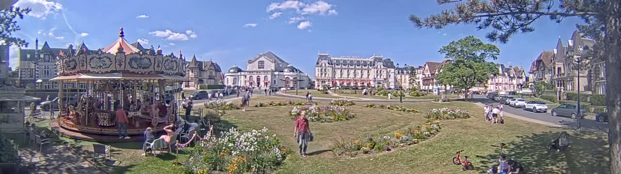 A carousel sits in a grassy town square on a sunny day, with several buildings and people visible in the background.