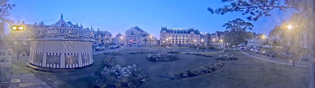 A carousel sits in a town square at dusk, next to several buildings including a large hotel, with cars parked along the streets and landscaped areas surrounding the square.