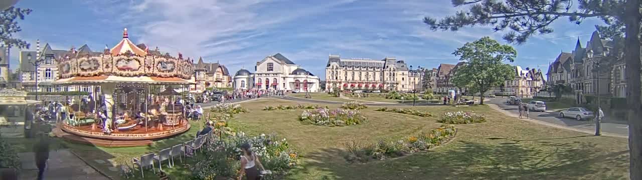 A carousel sits in a town square with several buildings, flowerbeds, and people, under sunny skies.