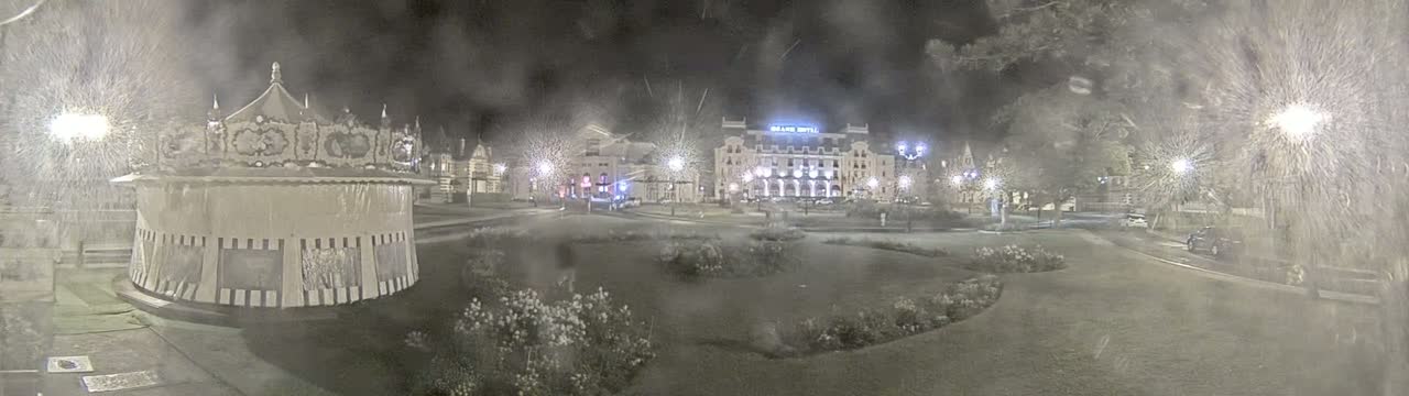 A nighttime scene of a snow-covered park with a carousel, buildings, and streetlights, under heavy snowfall.