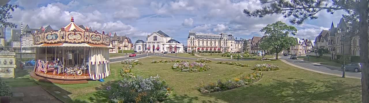 A carousel sits in a grassy town square, under a partly cloudy sky, near several buildings, some of which appear to be hotels.
