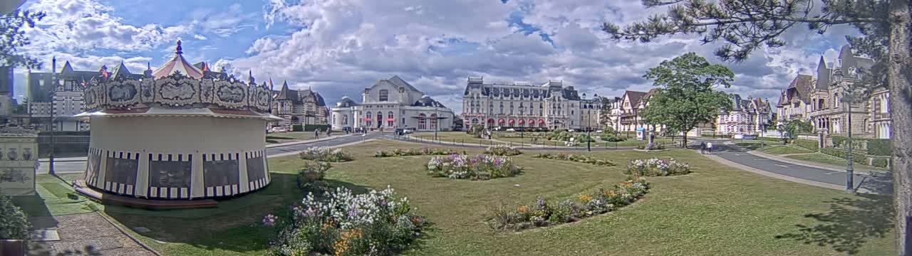 A carousel sits in a grassy town square in front of several buildings under a partly cloudy sky.