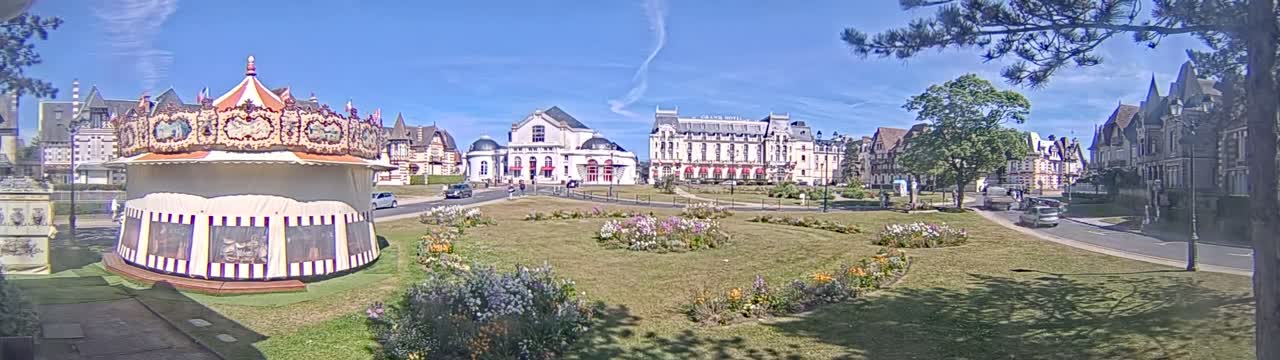 A carousel sits in a grassy town square, surrounded by several buildings under a sunny sky.
