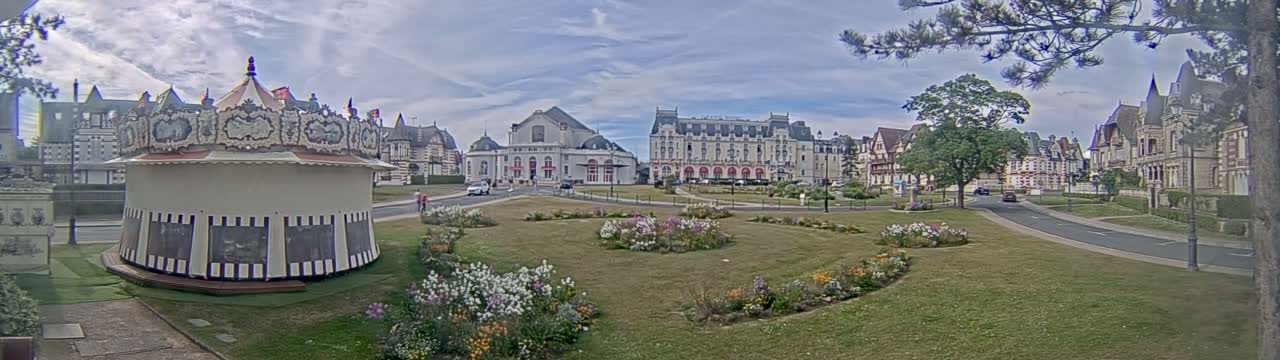 A carousel sits in a grassy town square bordered by flowerbeds, in front of several large buildings under a partly cloudy sky.