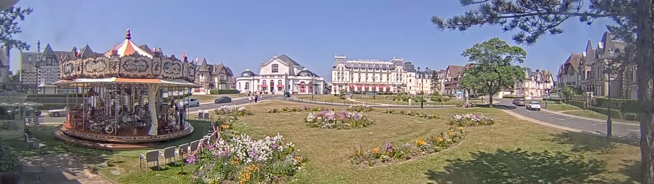 A carousel sits in a town square on a sunny day, surrounded by buildings, flowerbeds, and trees.