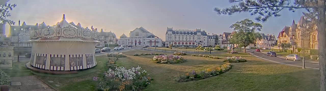 A carousel sits in a grassy park area in front of several large buildings under a clear, sunny sky.