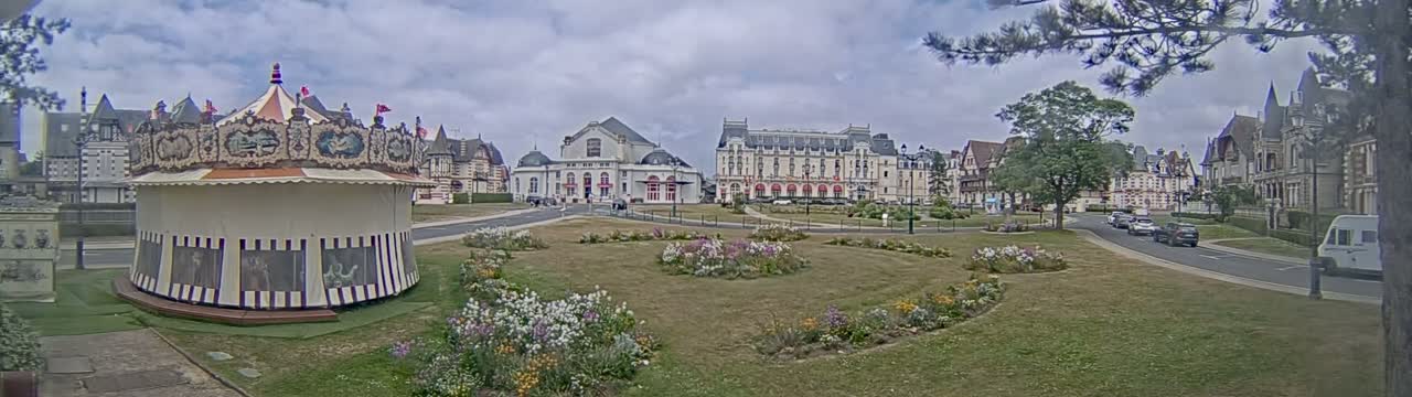 A carousel sits in a grassy town square, bordered by several large buildings under a partly cloudy sky.