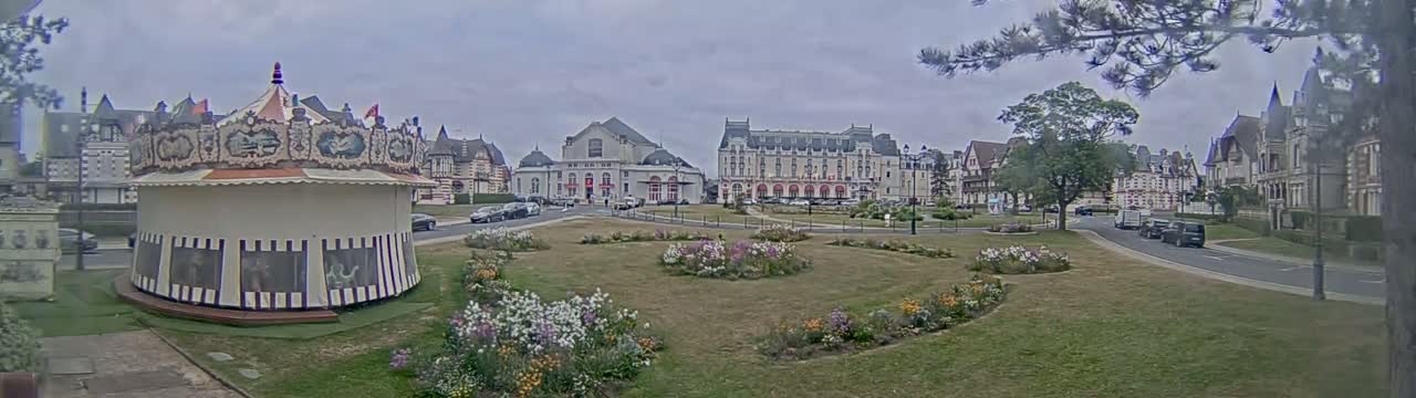 A carousel sits in a town square with flower gardens and several buildings, under an overcast sky.