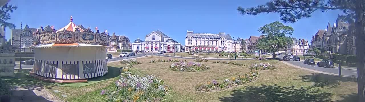 A carousel sits in a town square on a sunny day, with several buildings, including a large hotel, visible in the background.