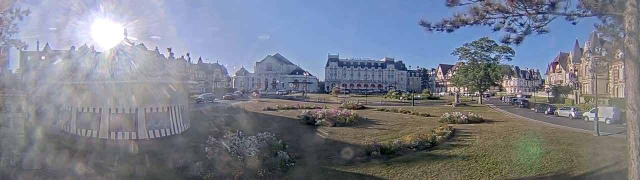 A sunny day reveals a town square with several large buildings, flowerbeds, and cars parked along a street.