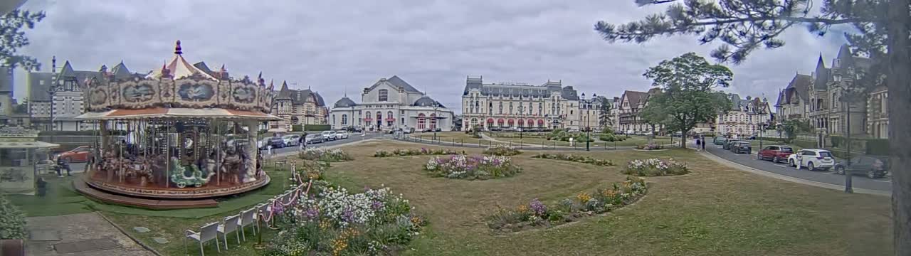 A carousel sits in a town square, surrounded by European-style buildings and flower gardens under an overcast sky.