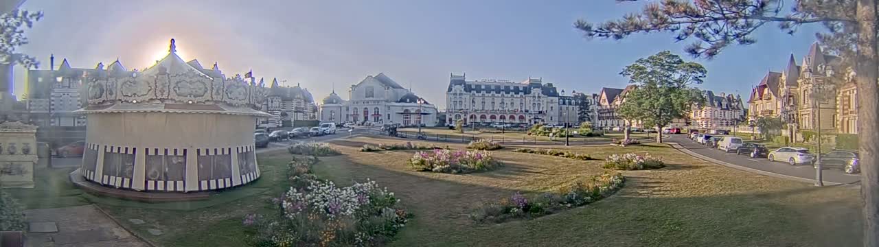 A carousel sits in a grassy park in front of several large buildings, on a sunny day.