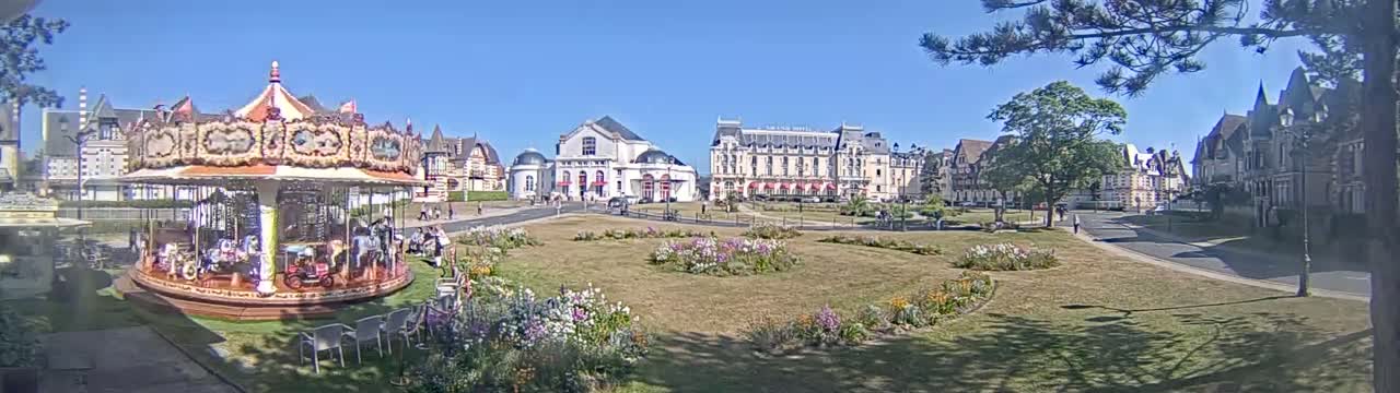 A carousel sits in a town square on a sunny day, surrounded by buildings and flower gardens.