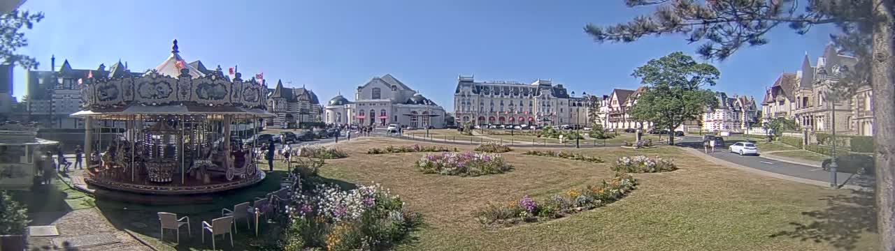 A carousel sits in a town square on a sunny day, with several buildings and flower gardens surrounding it.