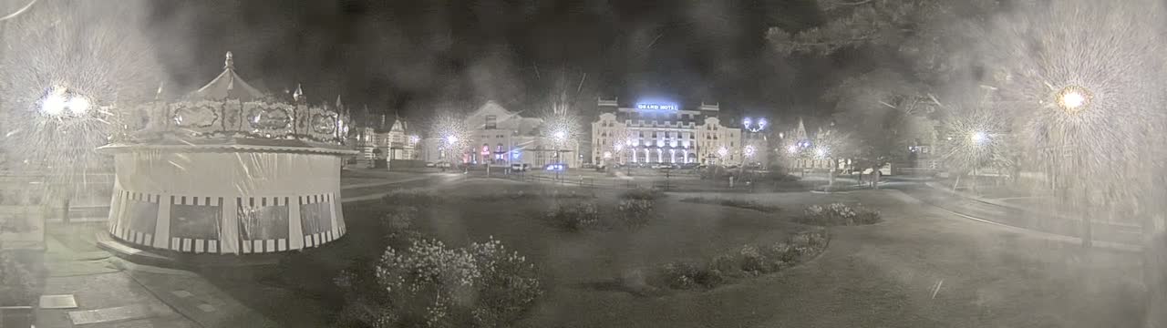 A snow-covered carousel sits in a park at night, with illuminated buildings and trees in the background.