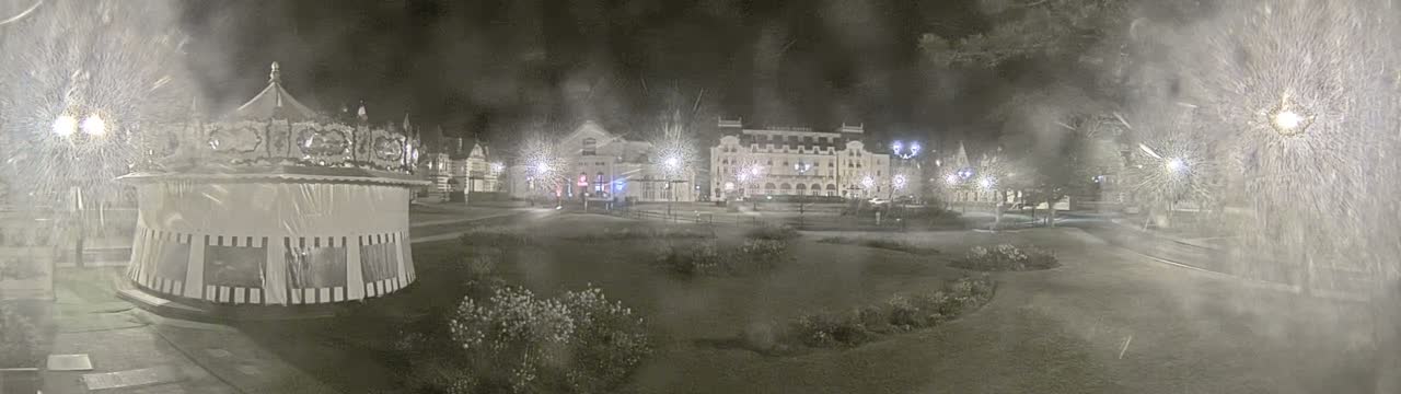 A carousel sits in a snow-covered town square at night, in front of a large building.