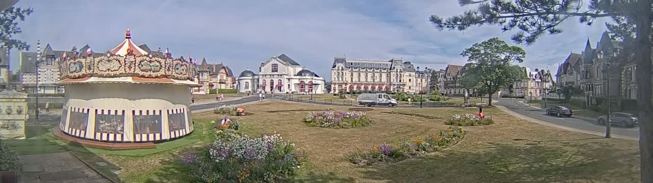 A carousel sits in a grassy town square under a partly sunny sky, surrounded by several buildings, flower gardens, and a few cars on the road.