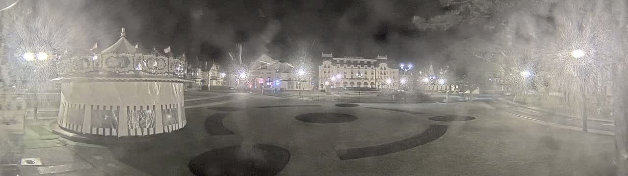 A snow-covered town square at night features a carousel, several buildings, and streetlights.
